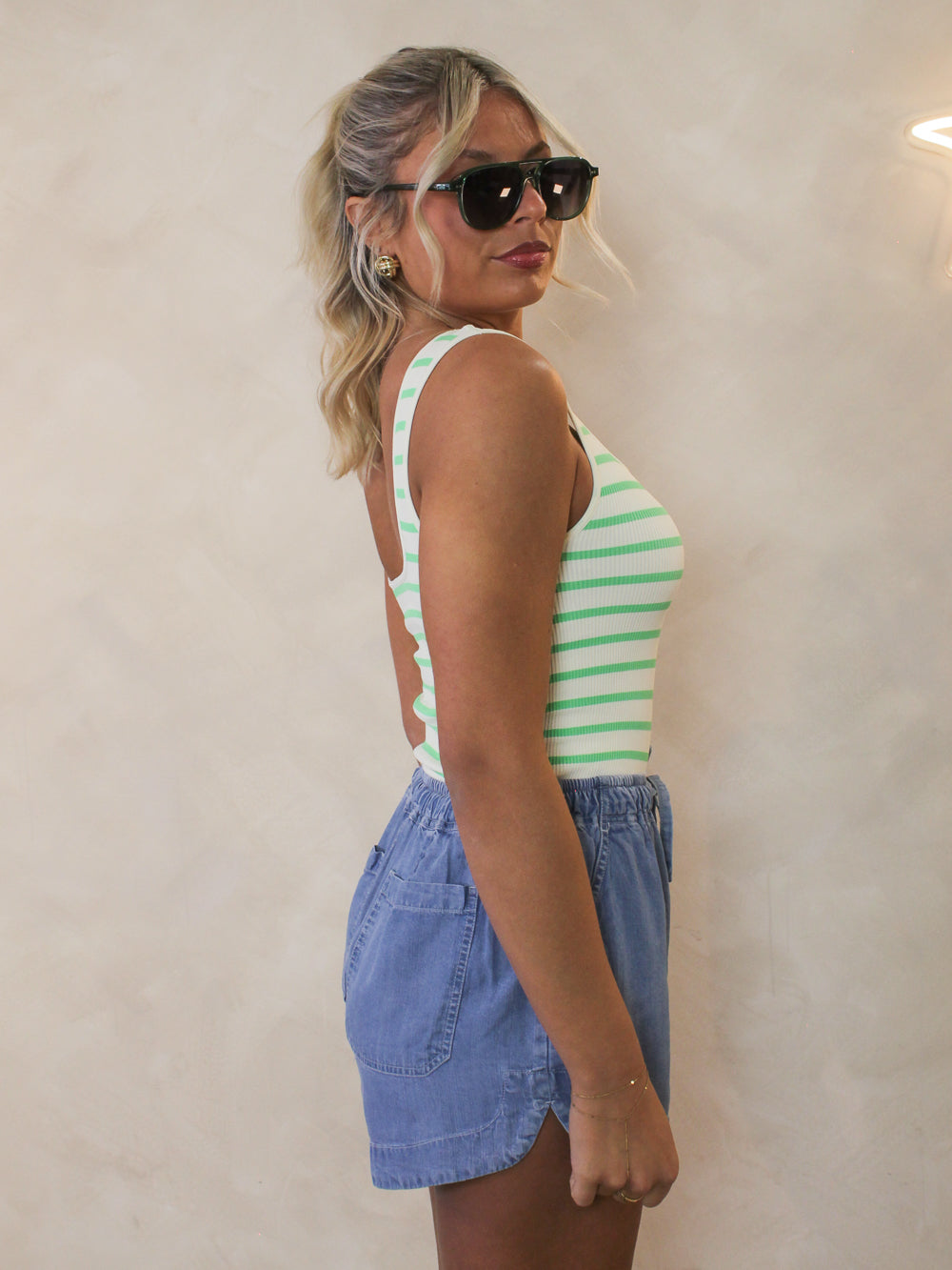 Model showing the side view of a green and white stripe bodysuit styled with denim shorts and sunglasses