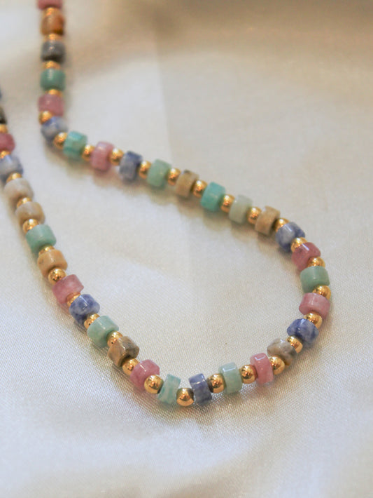 A detail view of beaded necklace featuring alternating pastel colored stone cubes in pink, green, blue, and beige, separated by small metallic gold spacer beads.