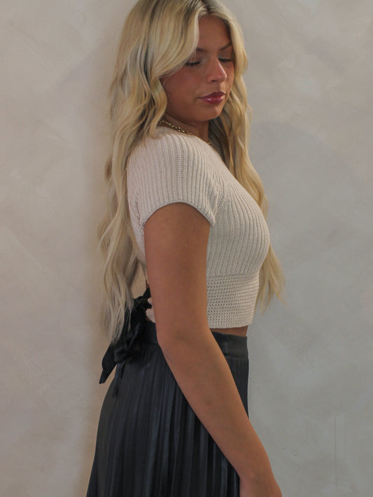 Woman wearing a beige knit, short sleeve sweater with a bow that ties in the back