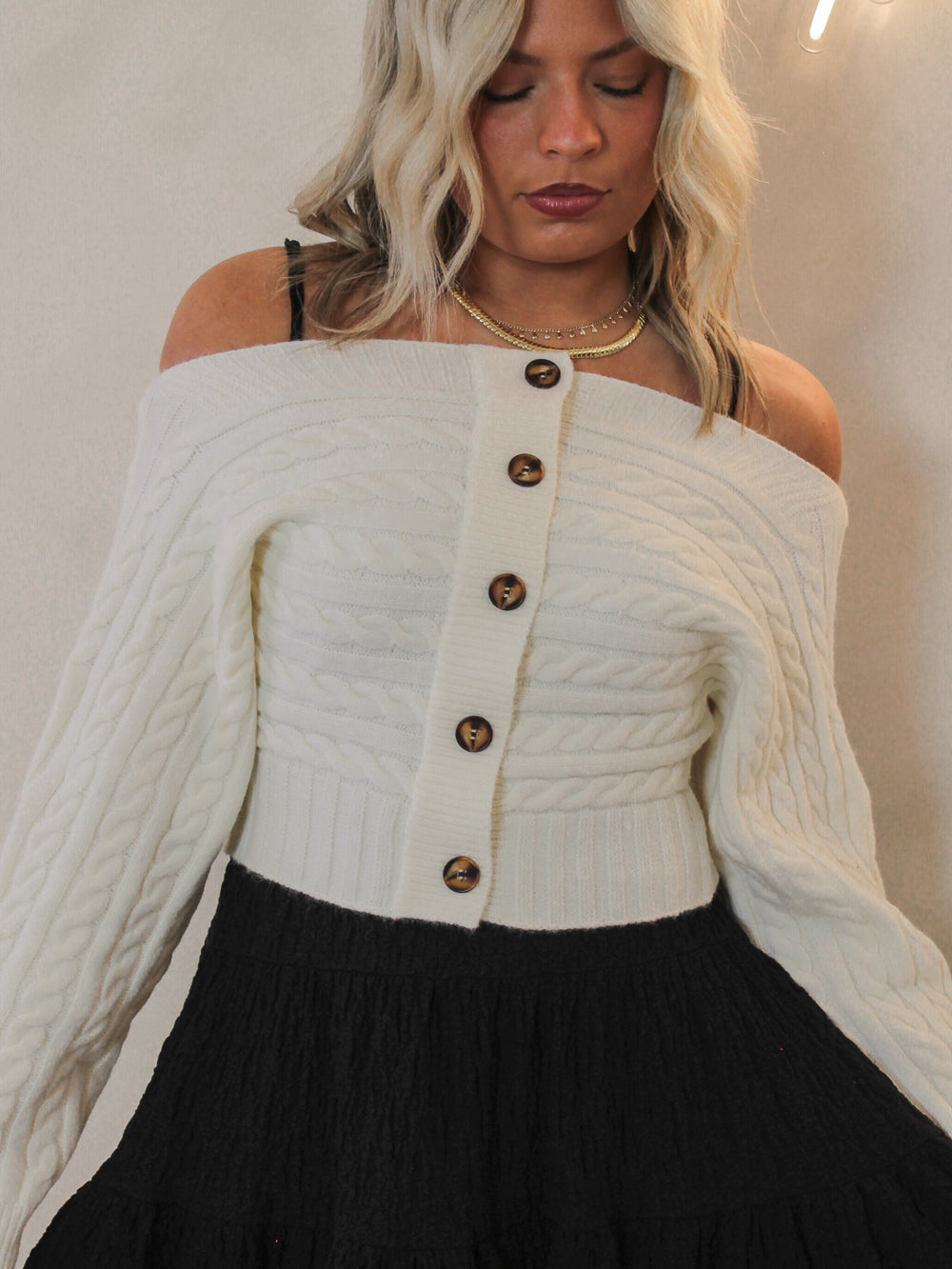 Woman wearing a white off-shoulder cable knit sweater with buttons and a black skirt.