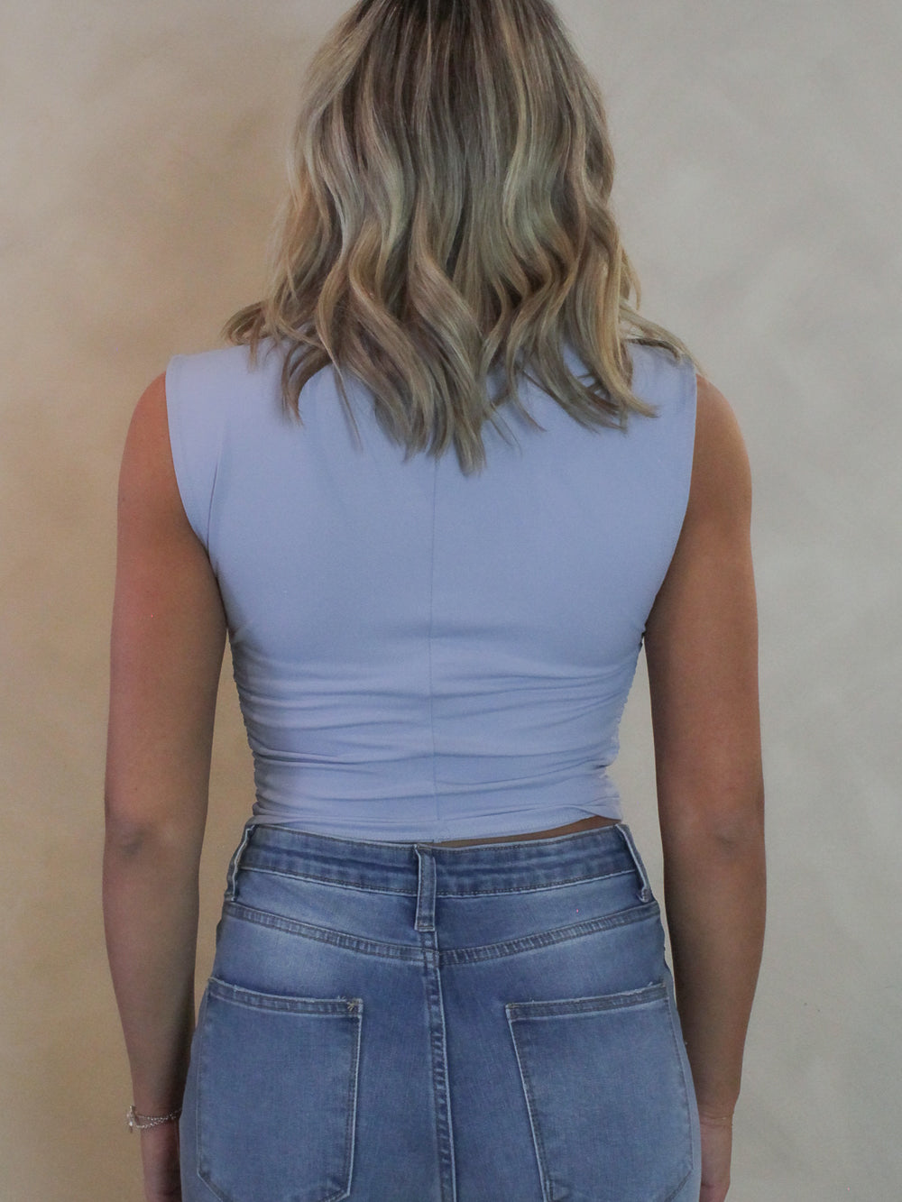 Back view of a woman wearing a baby blue sleeveless top with ruched sides and mock neck.