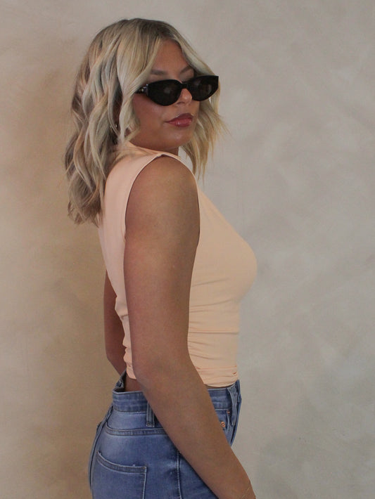 Woman wearing a coral sleeveless top with mock neck and ruched sides, sunglasses and denim pants