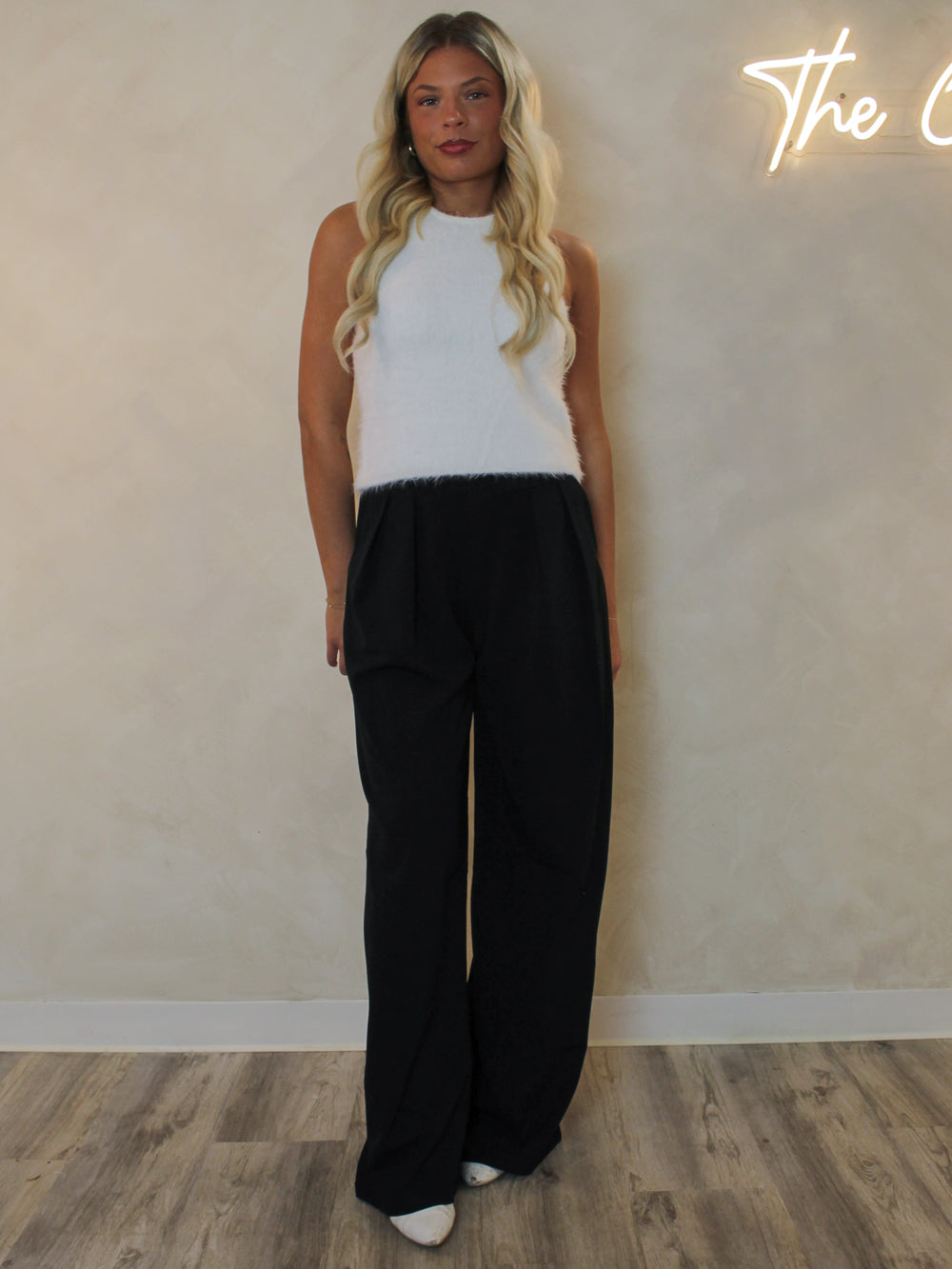 Woman wearing a white fuzzy knit tank with a high neckline and styled with black trousers and white boots.