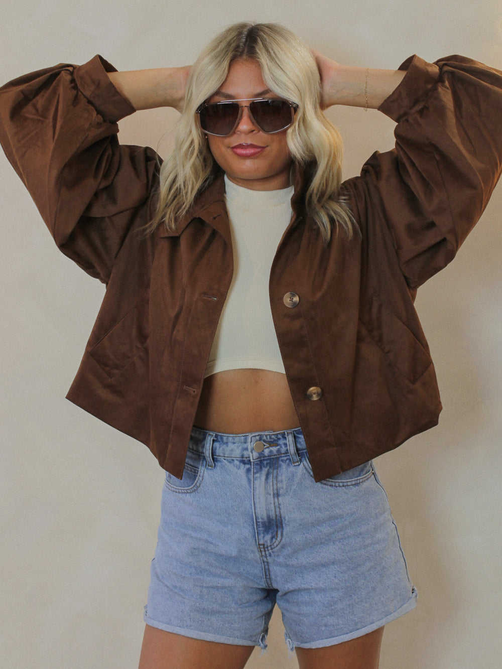Person wearing a brown Steve Madden jacket, white top, and blue shorts with sunglasses against