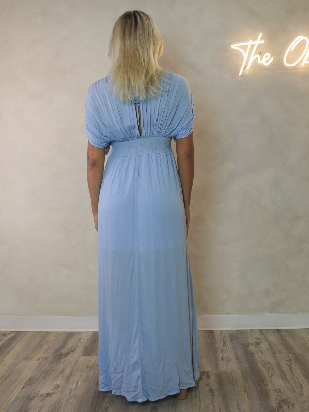 A woman viewed from behind wearing a light blue maxi dress with short drape sleeve and a smocked waist