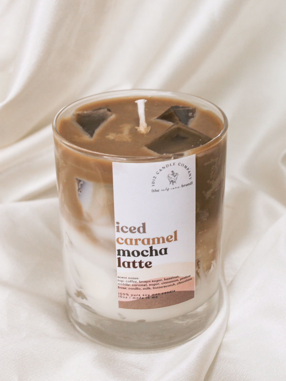 A candle labeled Iced Caramel Mocha Latte in a clear jar that looks just like an iced latte with realistic layered look and ice cubes