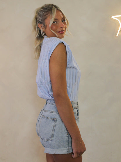 Woman wearing a sleeveless light blue button up tucked into denim shorts