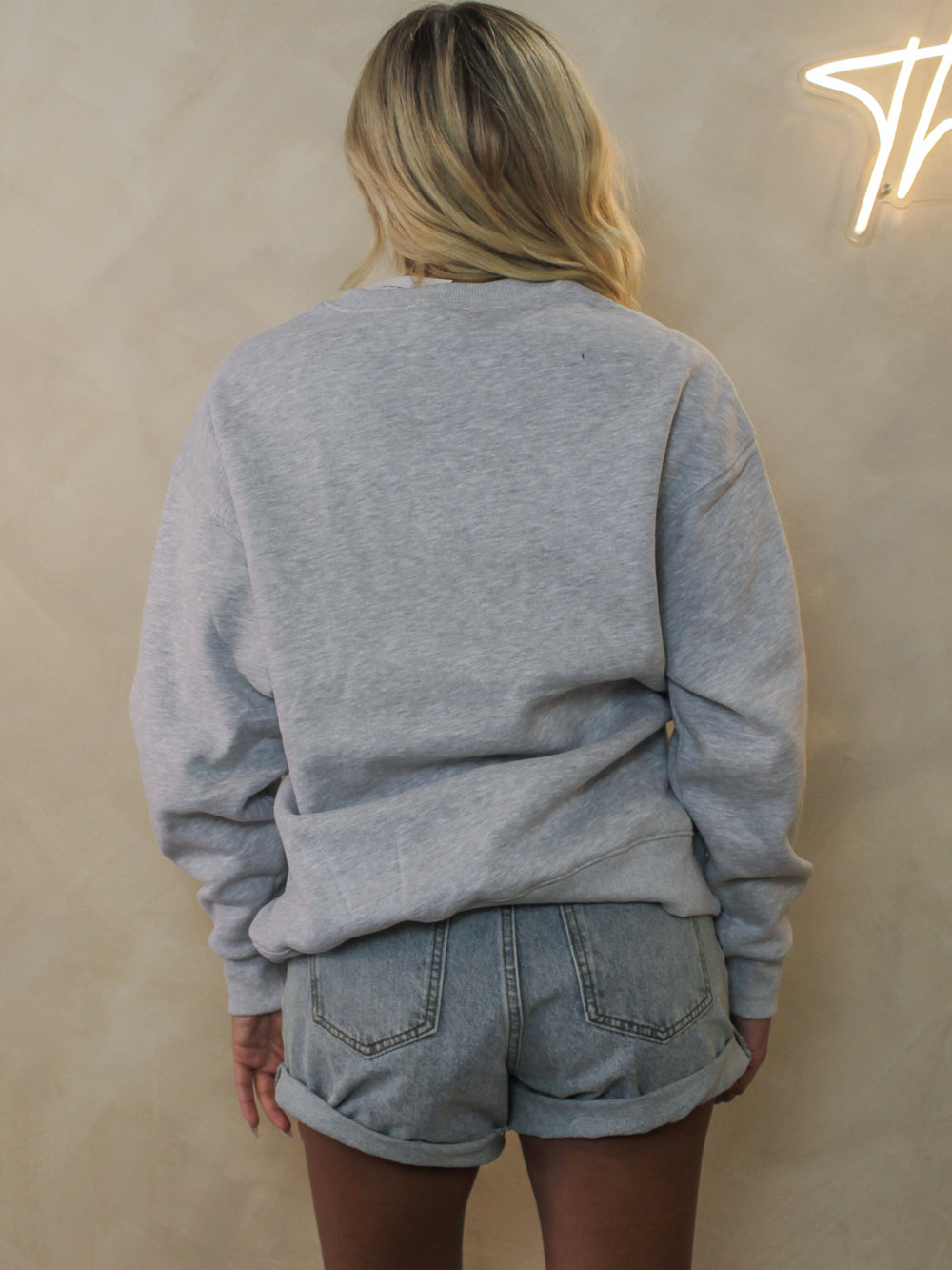 A woman viewed from behind wearing a light heather gray crewneck sweatshirt and light-wash denim short.