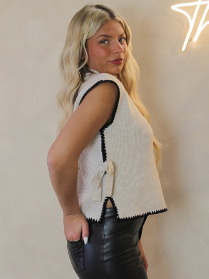 Woman wearing a cream sleeveless sewater with black trim and bow accents 