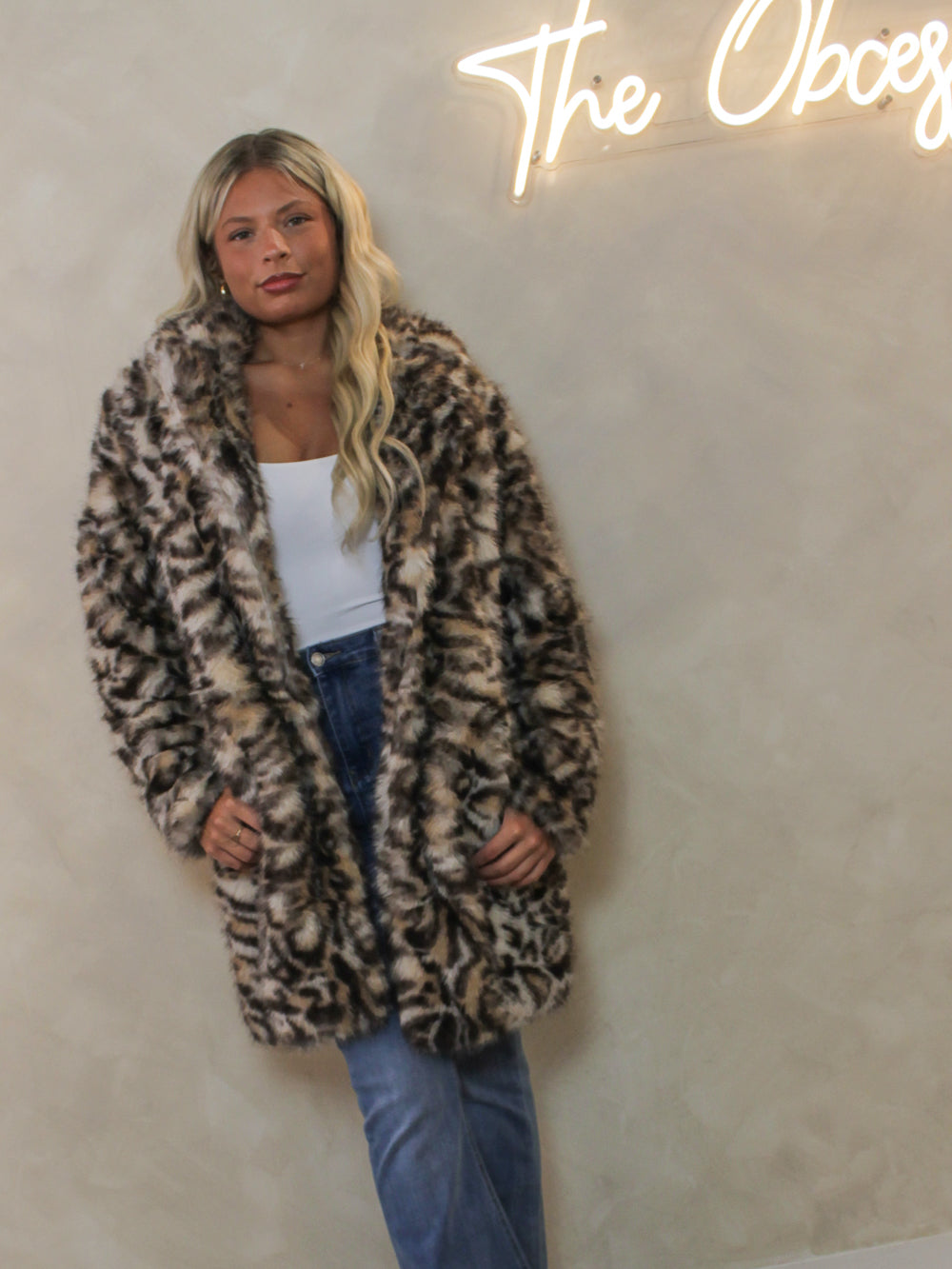 A woman wearing a faux fur coat with a  leopard print pattern, styled with a white tank top and blue jeans.