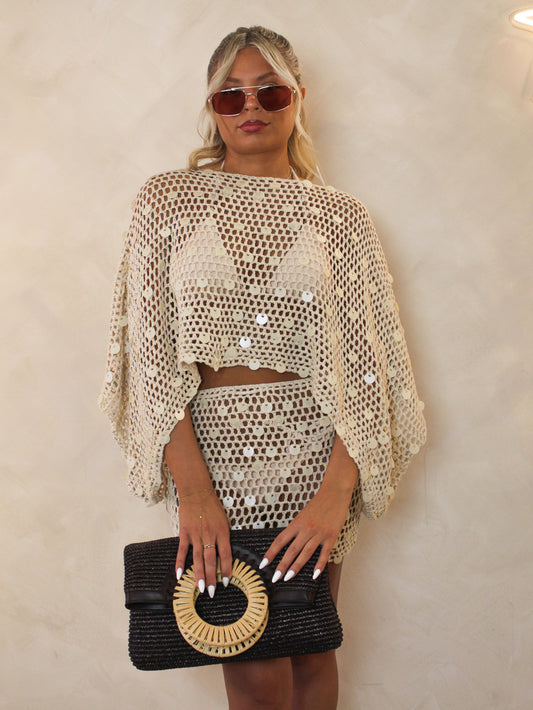 Woman wearing a beige crochet top and skirt set with sunglasses and a black clutch.