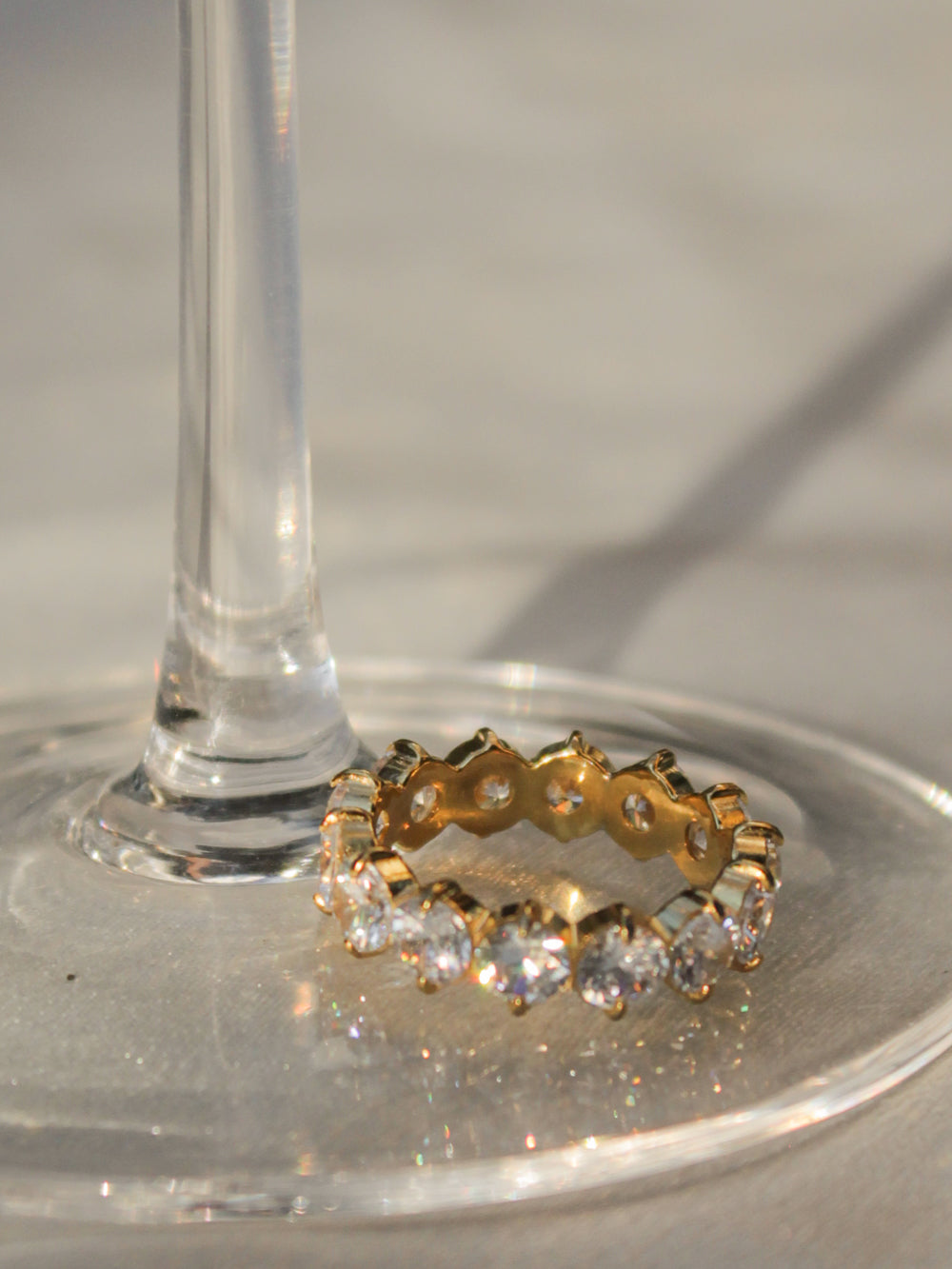A gold eternity ring with clear gemstones, resting on the base of a clear glass stem