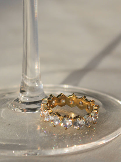 A gold eternity ring with clear gemstones, resting on the base of a clear glass stem