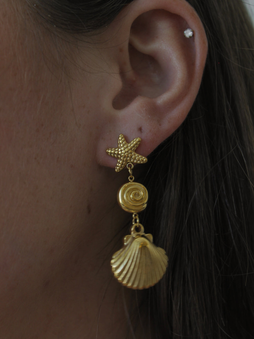 A close-up of a girl wearing a gold drop earring with a textured starfish stud, a spiral shell charm, and a larger shell that dangles