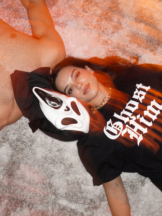 Halloween aesthetic picture with a woman wearing a "Ghost Him" Tee laying next to a person wearing a ghost face mask