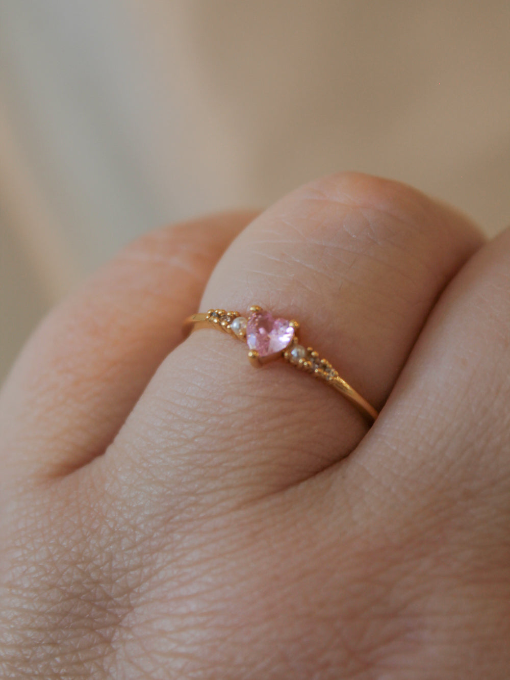 A gold ring with a small heart-shaped pink gemstone and tiny accent stones, shown worn on a finger.