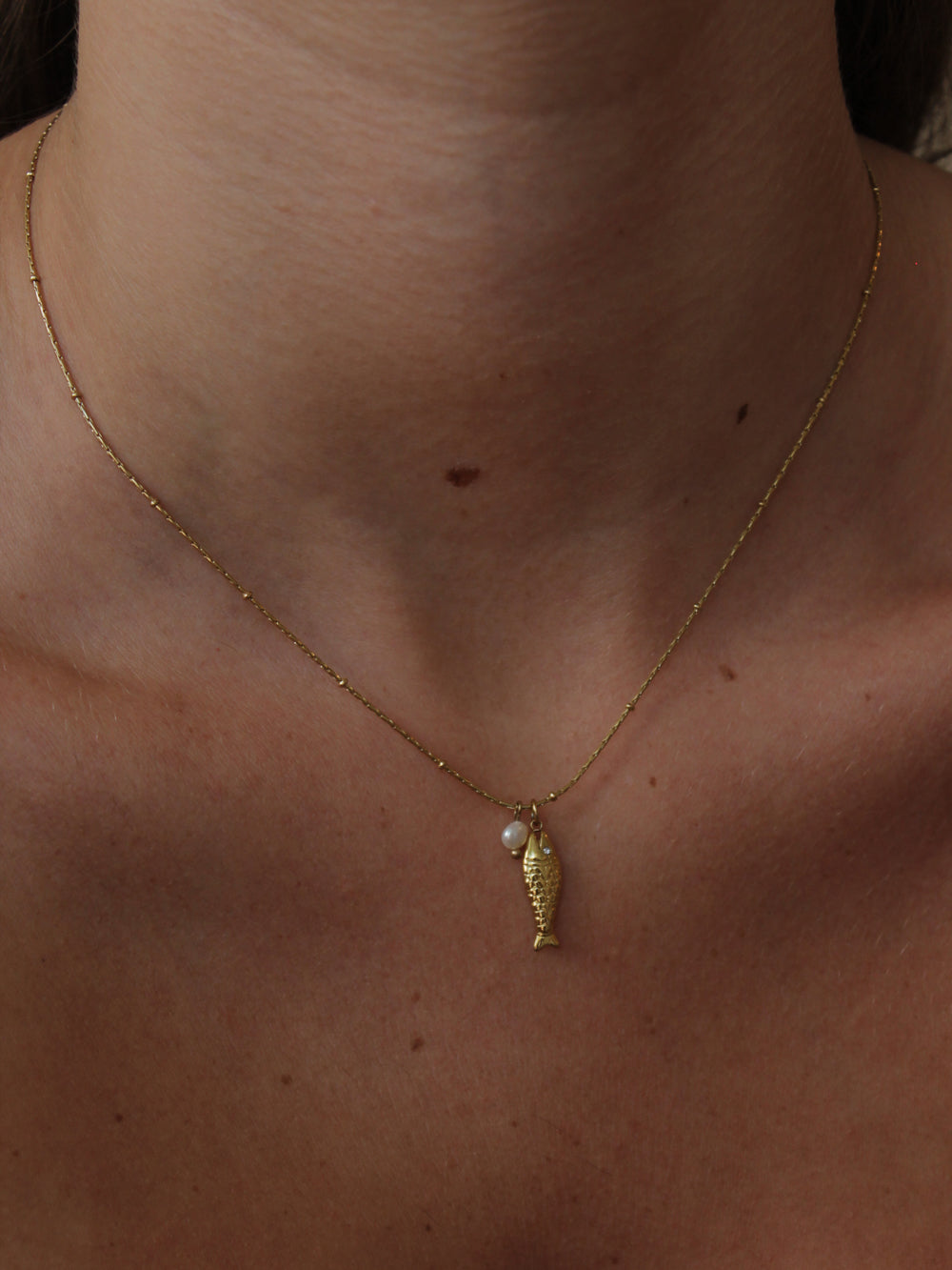 Dainty gold necklace with a fish and pearl charms on a model