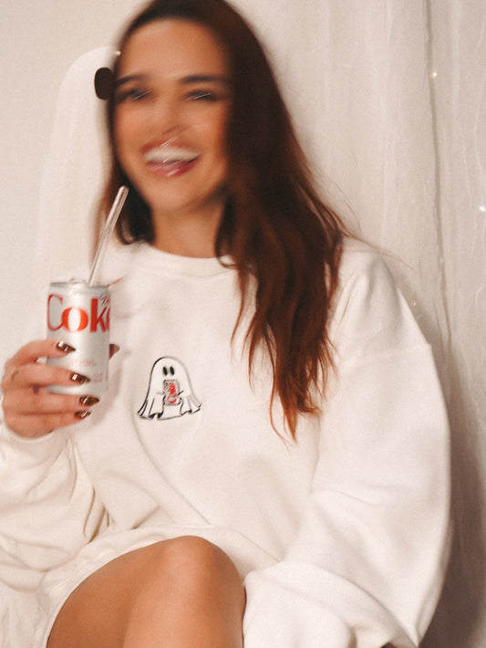 Woman wearing a white halloween theme sweatshirt with an embroidered ghost design, holding a can of diet coke.