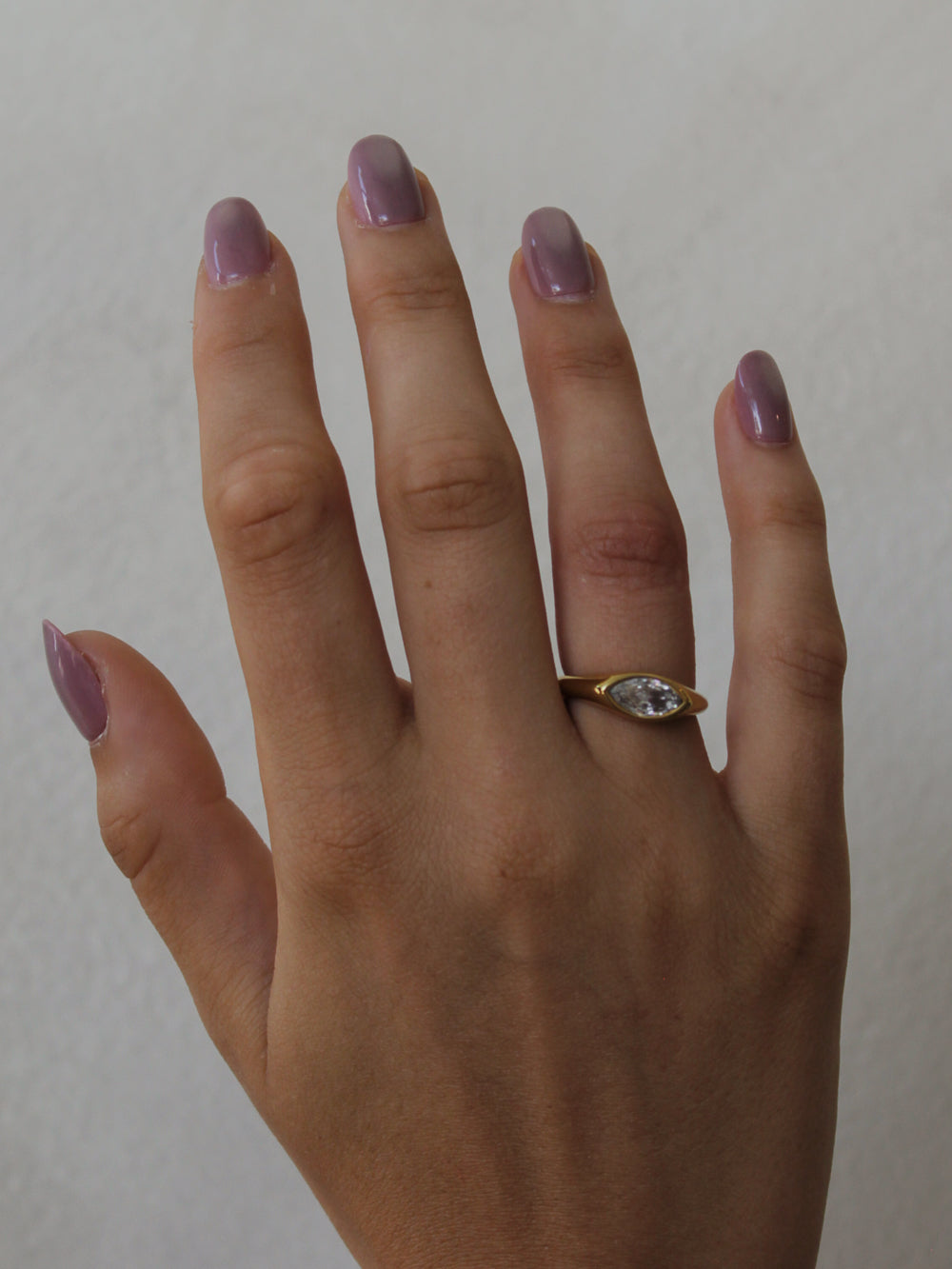 A close-up of a hand with lavender-painted nails wearing a gold ring with a marquise-cut clear gemstone.