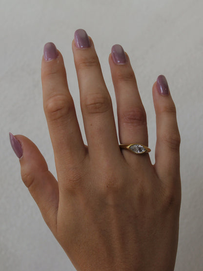 A close-up of a hand with lavender-painted nails wearing a gold ring with a marquise-cut clear gemstone.