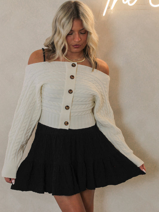 Woman wearing a white off-shoulder sweater with buttons over a black mini dress
