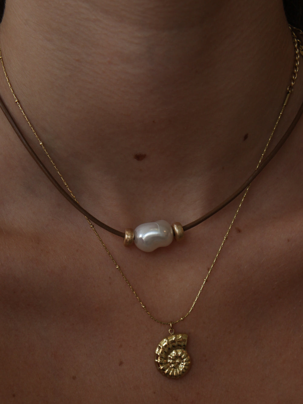 Close-up of a person wearing a gold necklace with a pearl pendant and another gold necklace shell charm