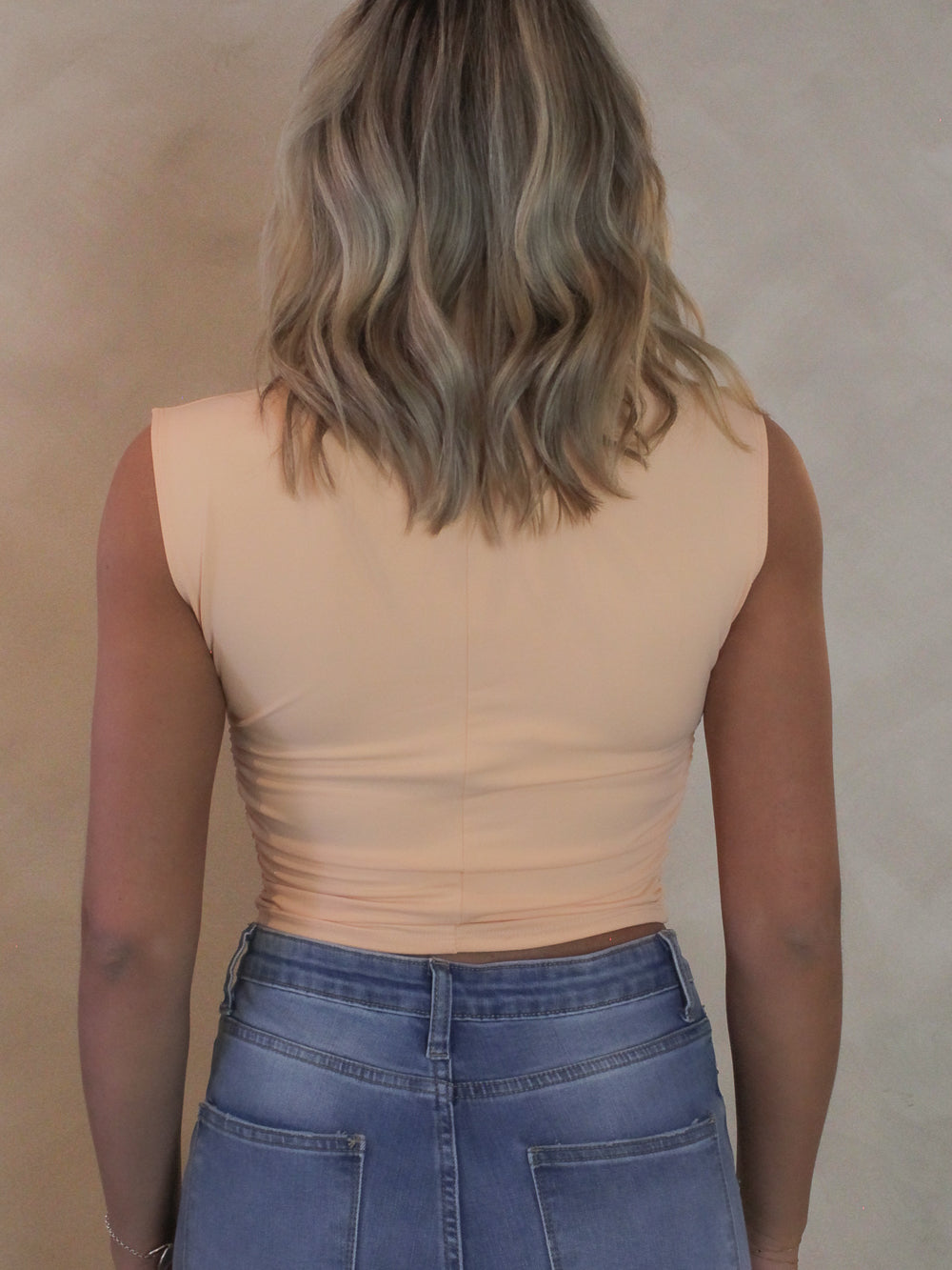 Back view of a woman wearing a light coral sleeveless top with mock neck, sunglasses and denim pants