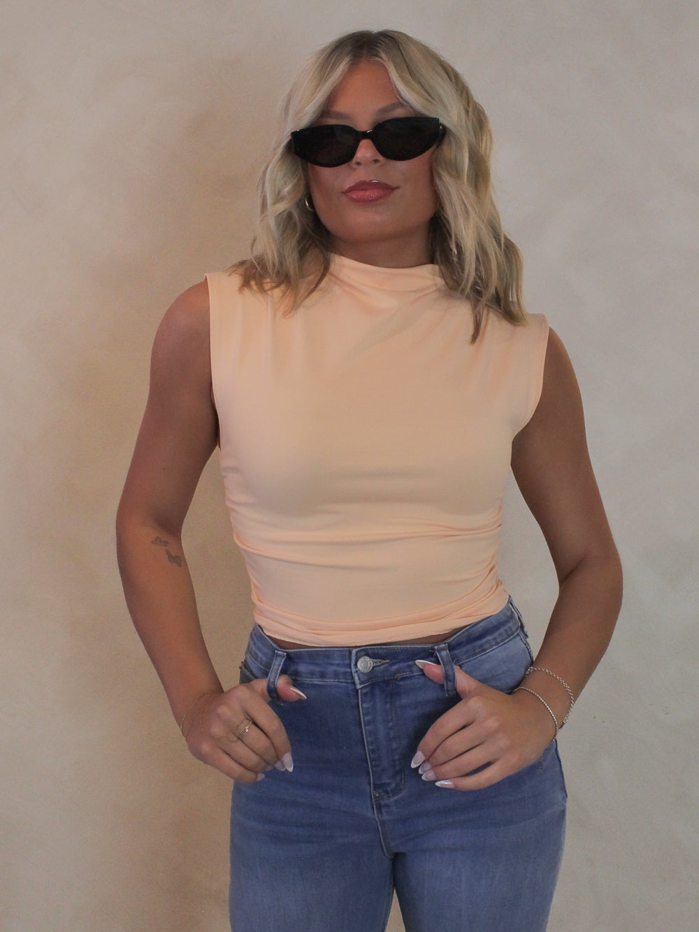 Woman wearing a light coral sleeveless top with mock neck, sunglasses and denim pants