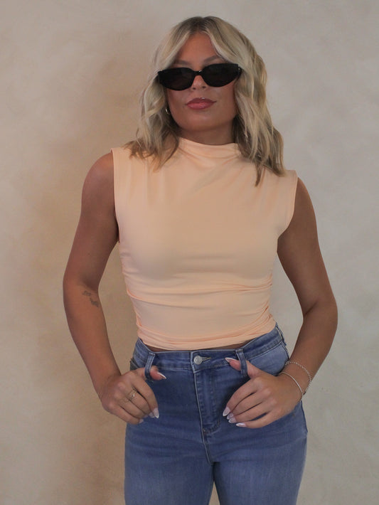 Woman wearing a light coral sleeveless top with mock neck, sunglasses and denim pants