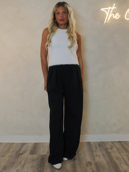Woman wearing a white fuzzy knit tank with a high neckline and styled with black trousers and white boots.