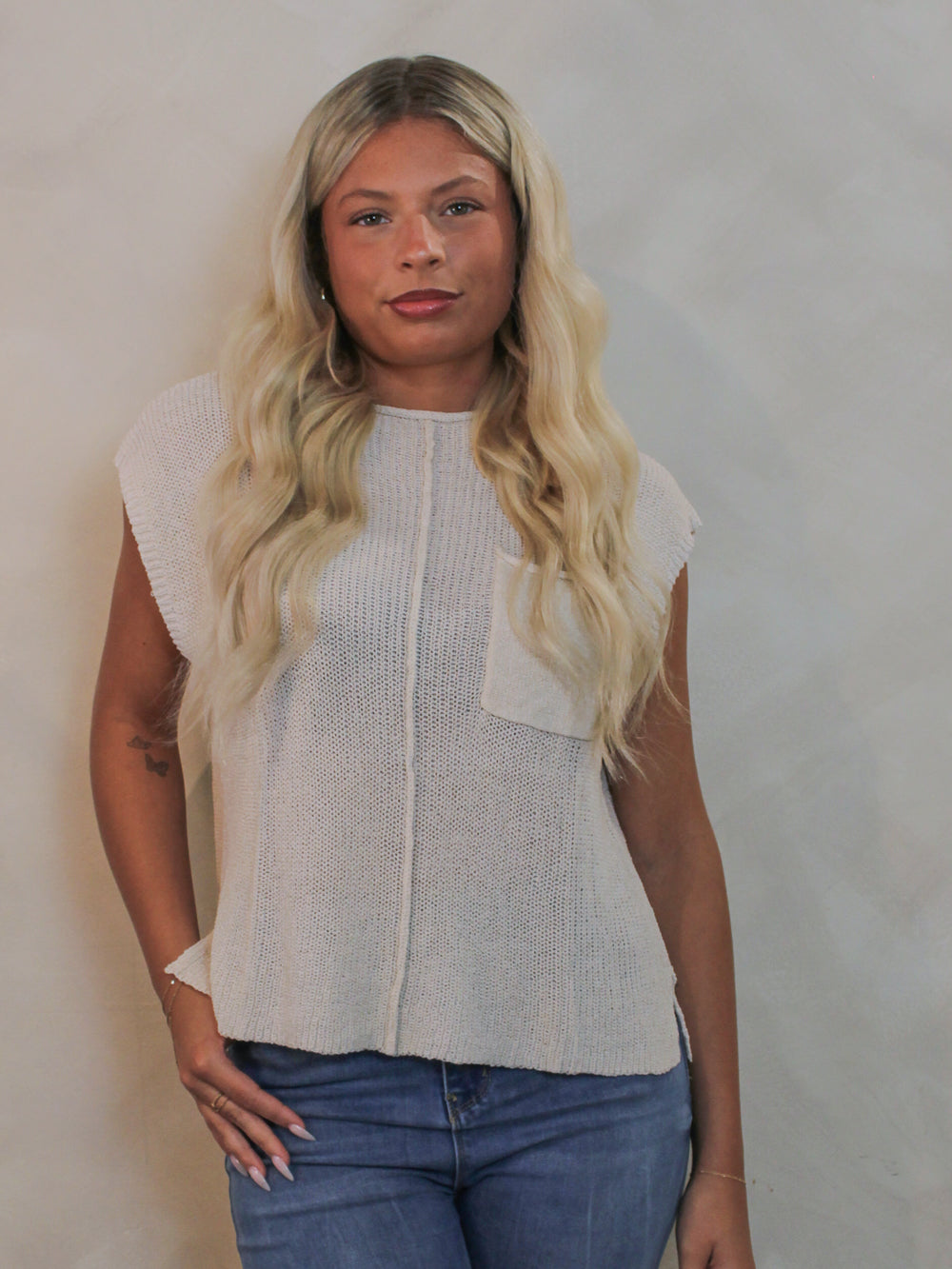 A woman wearing a sleeveless off-white knit top with a chest pocket and blue jeans