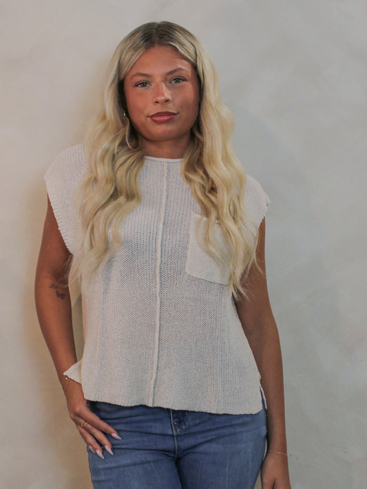 A woman wearing a sleeveless off-white knit top with a chest pocket and blue jeans