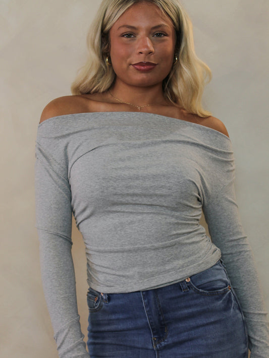A woman wearing a light gray off-the-shoulder long-sleeve top and blue jeans.