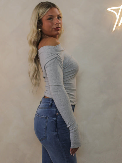 A woman wearing a light gray off-the-shoulder long-sleeve top and blue jeans. styled for an everyday outfit