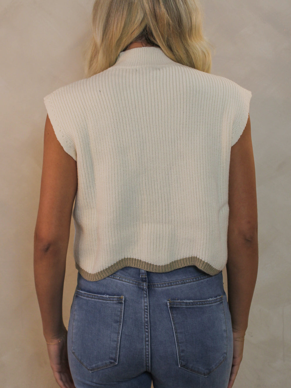 Back view of a woman wearing a cream, sleeveless sweater with a mock neck and scallop hem design