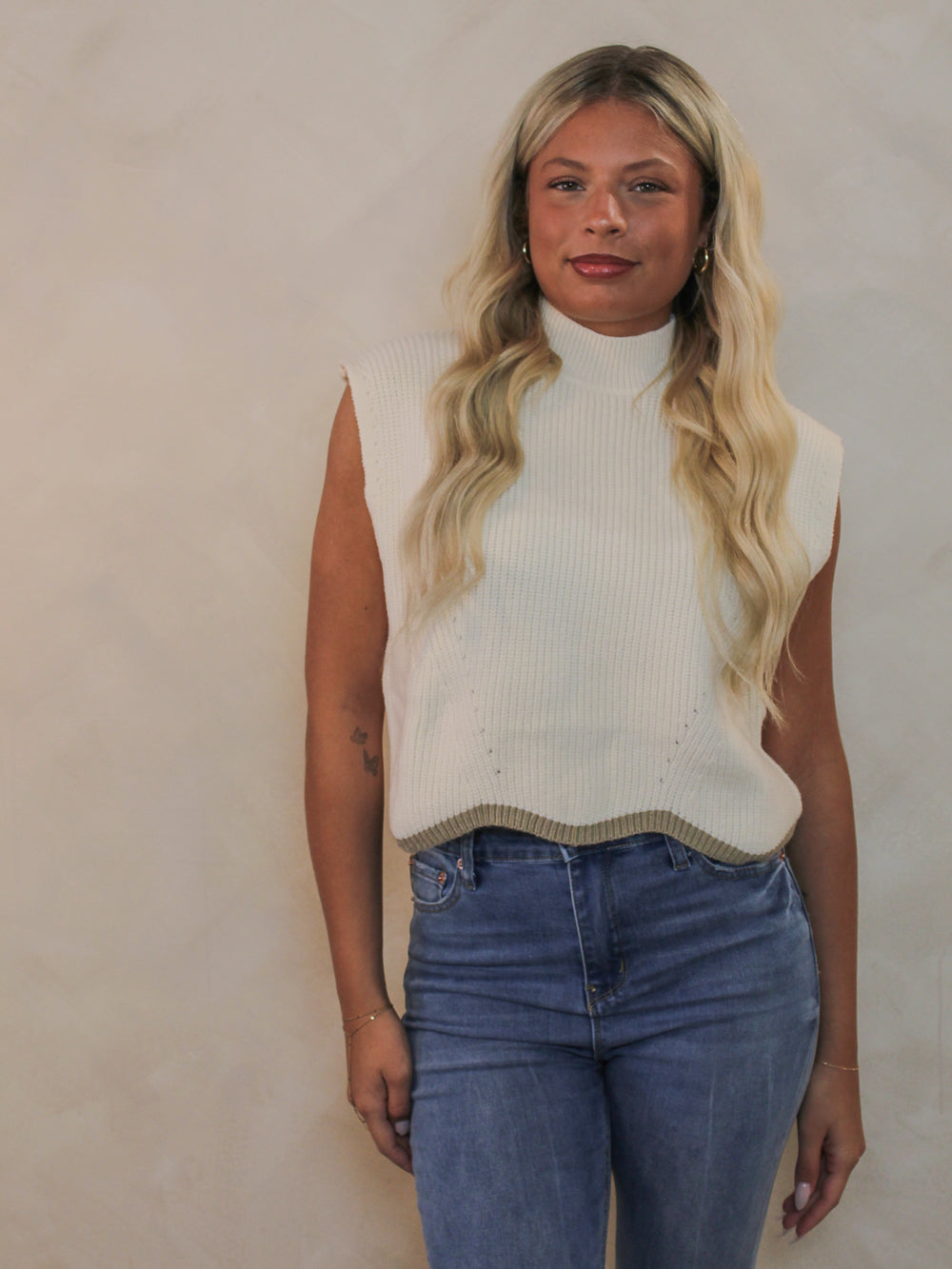 A woman wearing a ribbed cream, sleeveless mock neck sweater vest and blue jeans. 