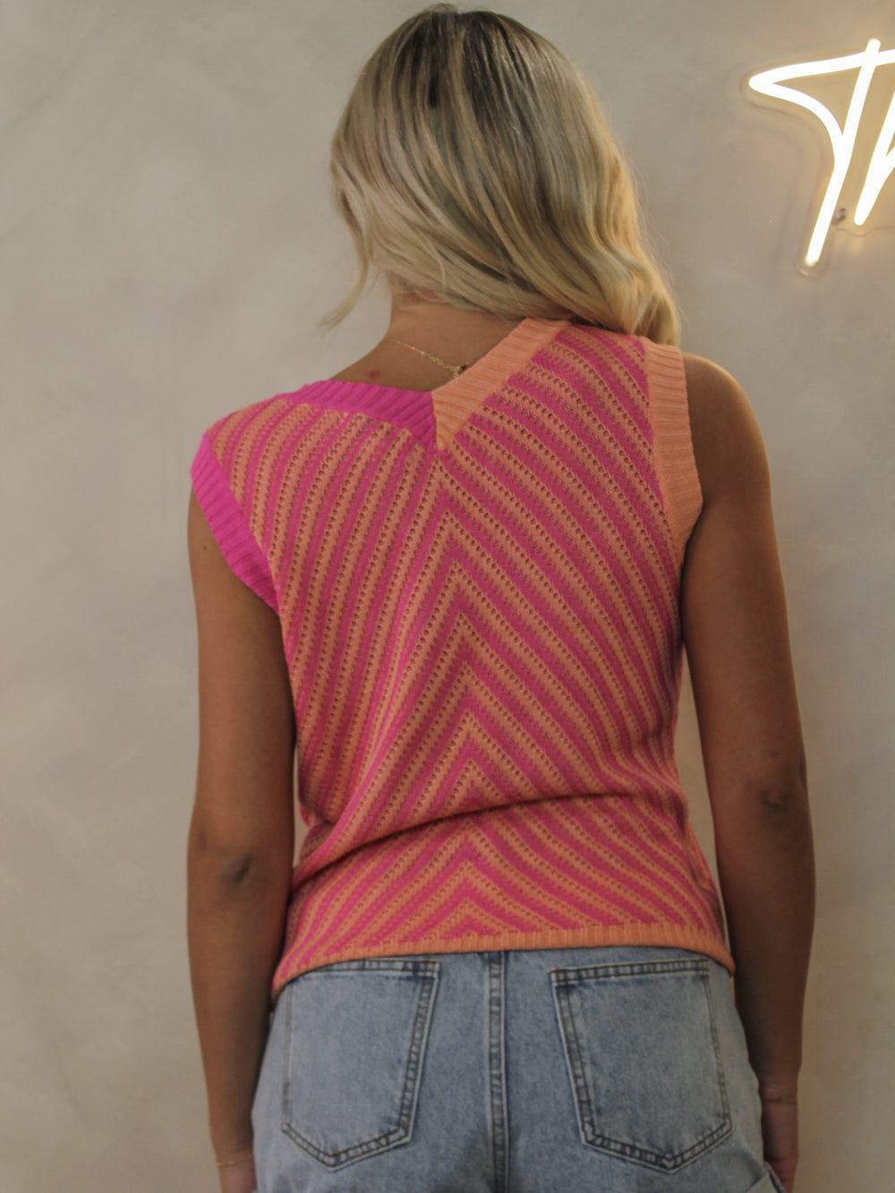 Back view of a model wearing a pink and orange striped sleeveless knit top styled with light wash denim.