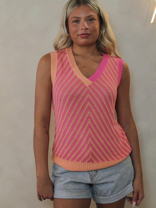 A model wearing a sleeveless knit sweater vest with a pink and peach chevron pattern, paired with denim shorts.