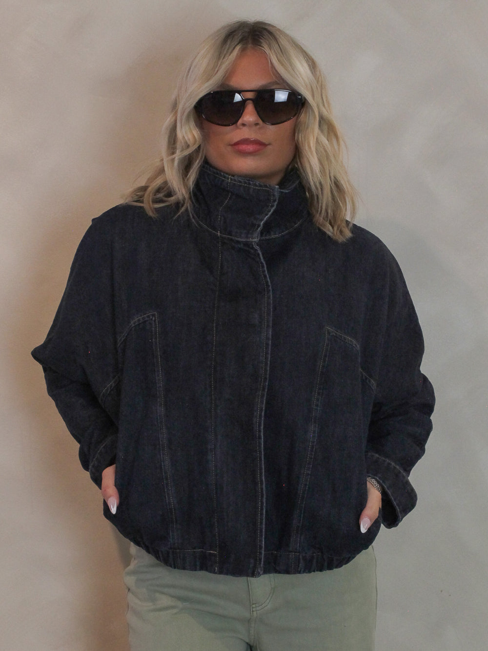 Woman wearing a dark denim, funnel neck jacket with sunglasses and sage green pants
