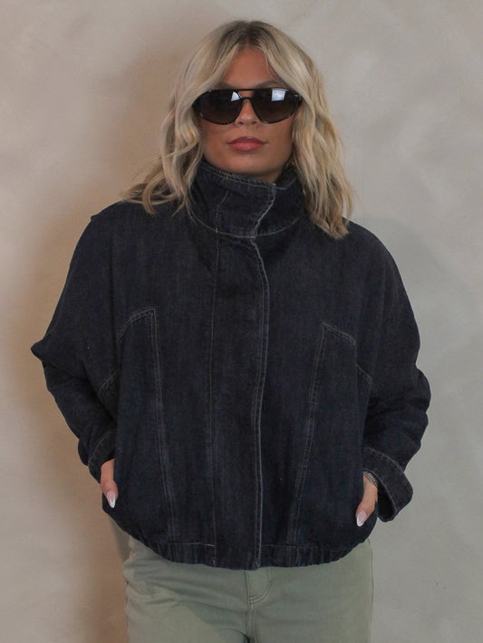 Woman wearing a dark denim, funnel neck jacket with sunglasses and sage green pants