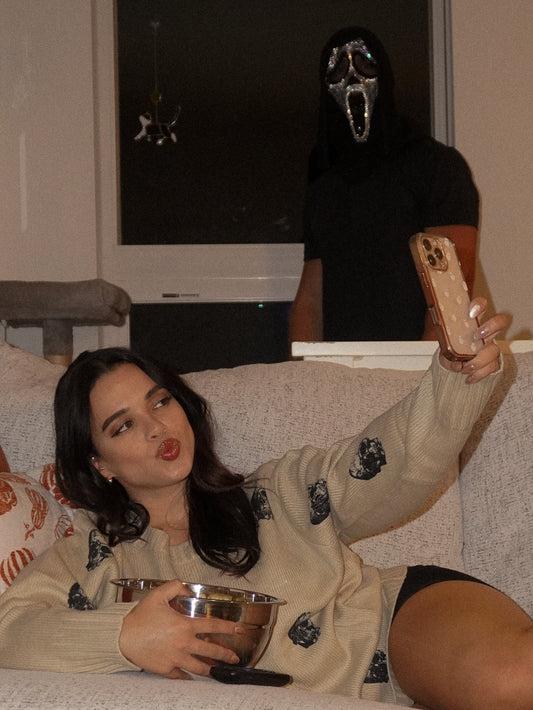 Woman wearing a skull print sweater laying on a couch with a man in a scream mask standing behind her for a halloween inspired photoshoot