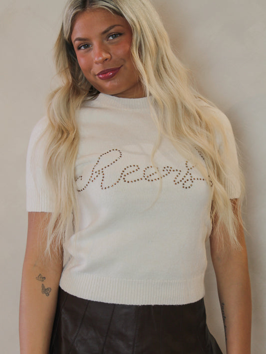 Ivory white sweater with cheers written across the front in rhinestones
