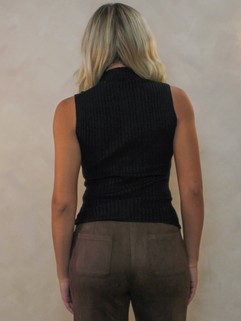 Model viewed from behind wearing a sleeveless black ribbed mock neck top and brown pants.