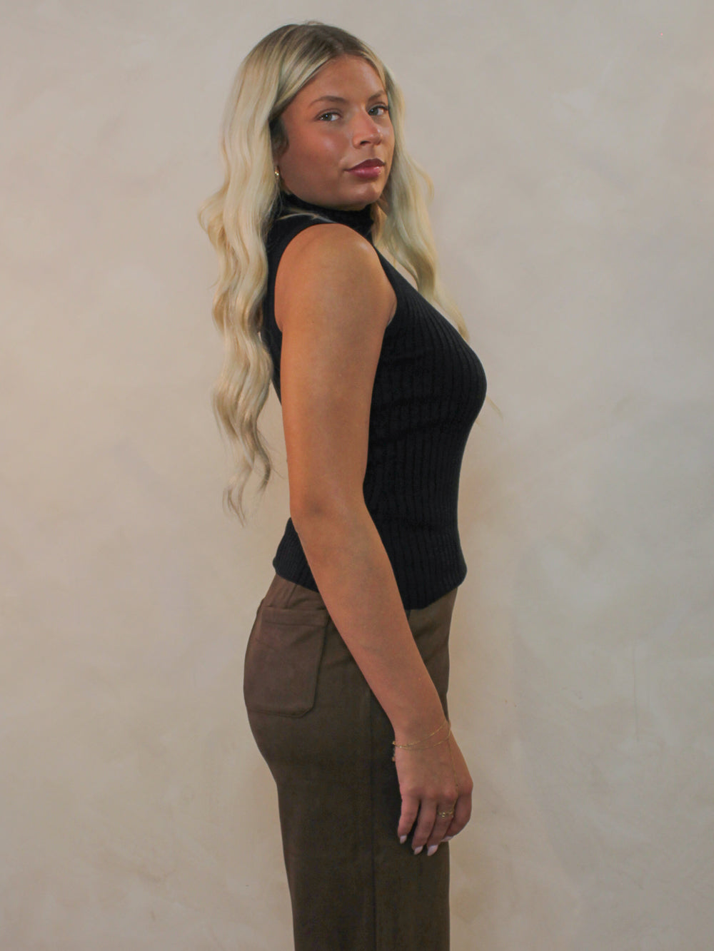 Model viewed from the side wearing a black ribbed sleeveless top and brown pants.