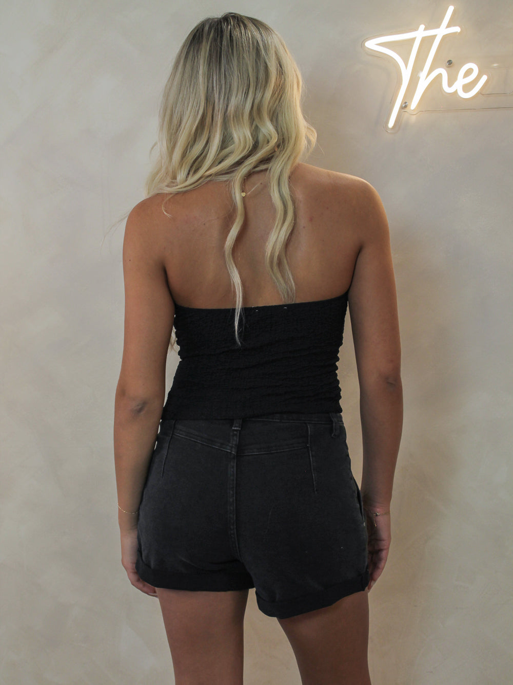 A woman viewed from behind wearing a black strapless tube top and black denim shorts.