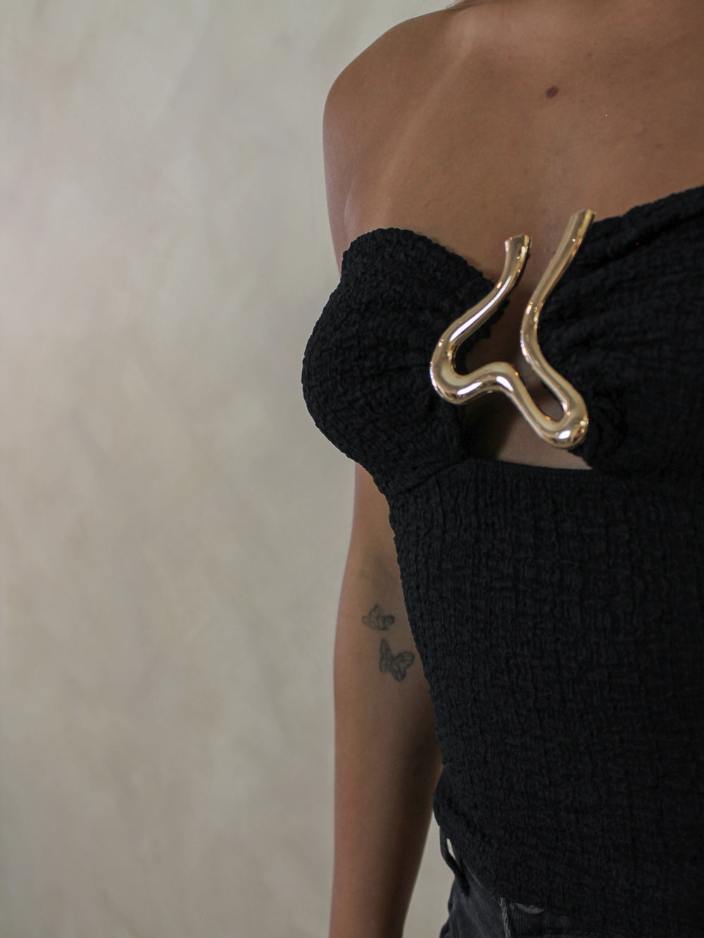 A close-up of a black textured strapless top with a prominent wavy metallic gold chest accent.