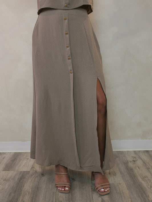 Model wearing a taupe-brown high-waisted maxi skirt with a gold front button-down detail and a side slit and styled with nude sandals