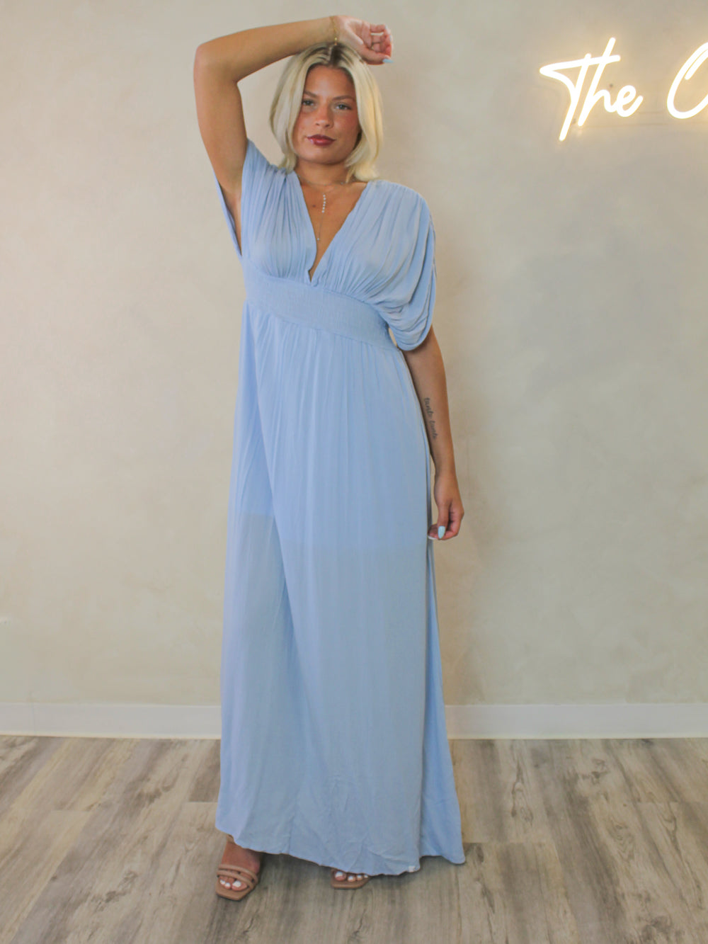 A woman modeling a light blue maxi dress with a deep V-neckline, draped short sleeves, and a flowing skirt.