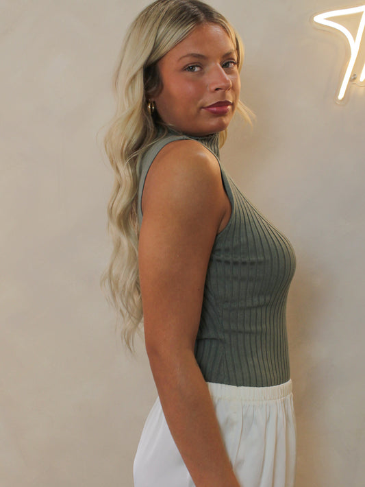Model shown from the side, wearing a ribbed sleeveless top in an olive green color paired with white bottoms.
