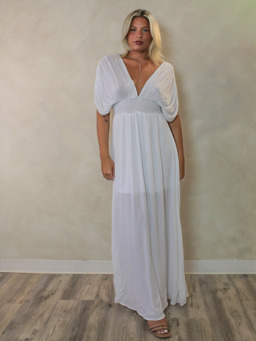 A woman modeling a white maxi dress with a deep V-neckline, short draping sleeves, and a flowing skirt.
